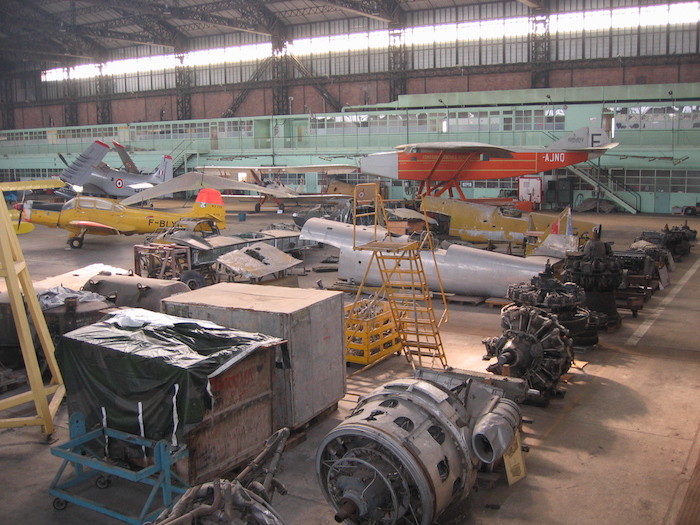 Une collection de moteurs de tous types et d'éléments de divers appareils est entreposée dans le fond de l'immense hangar.